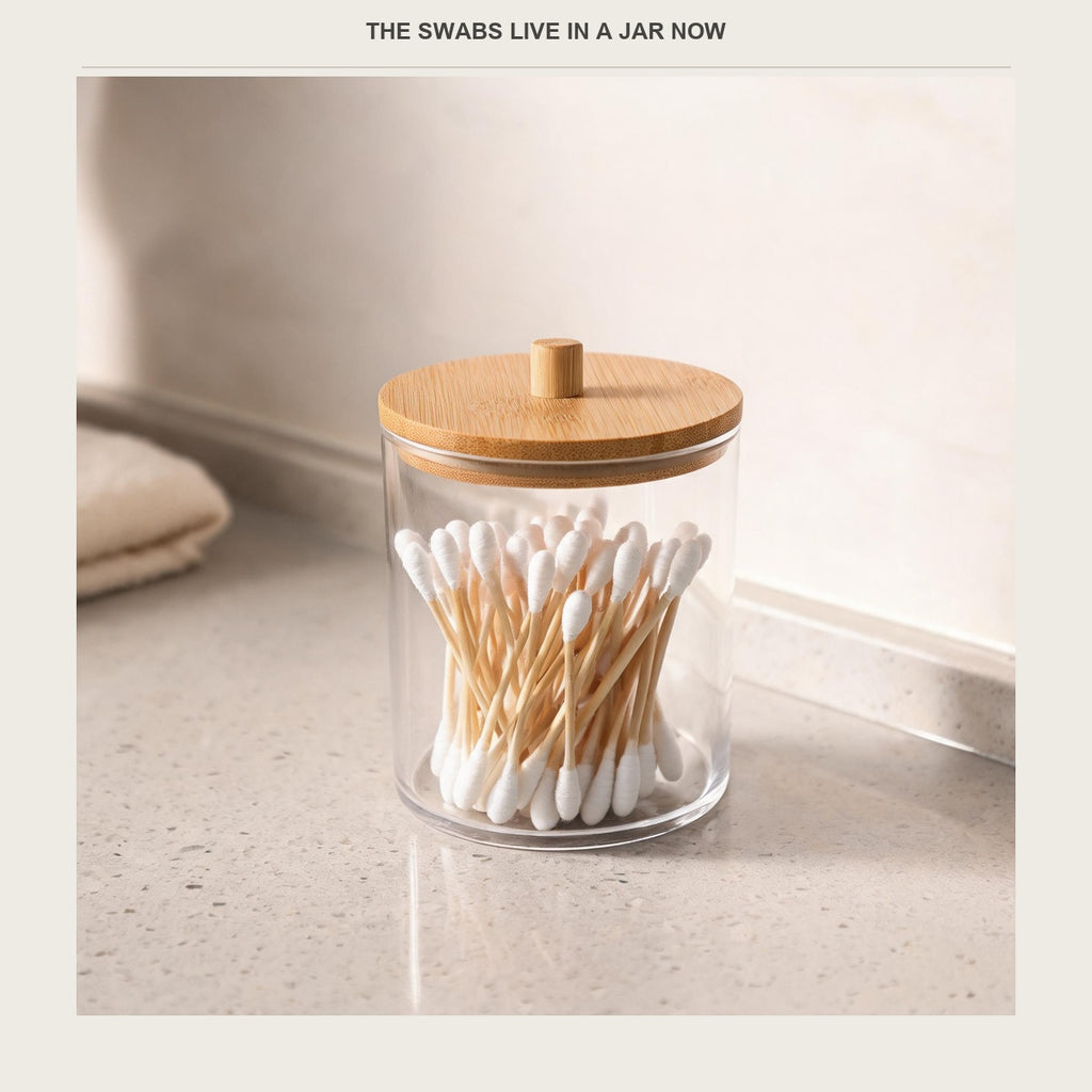 Clear acrylic cotton swab jar with bamboo lid on a pale bathroom counter, filled with swabs