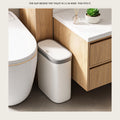 Slim white trash can with a grey rim standing in the narrow gap between a toilet and a wood vanity