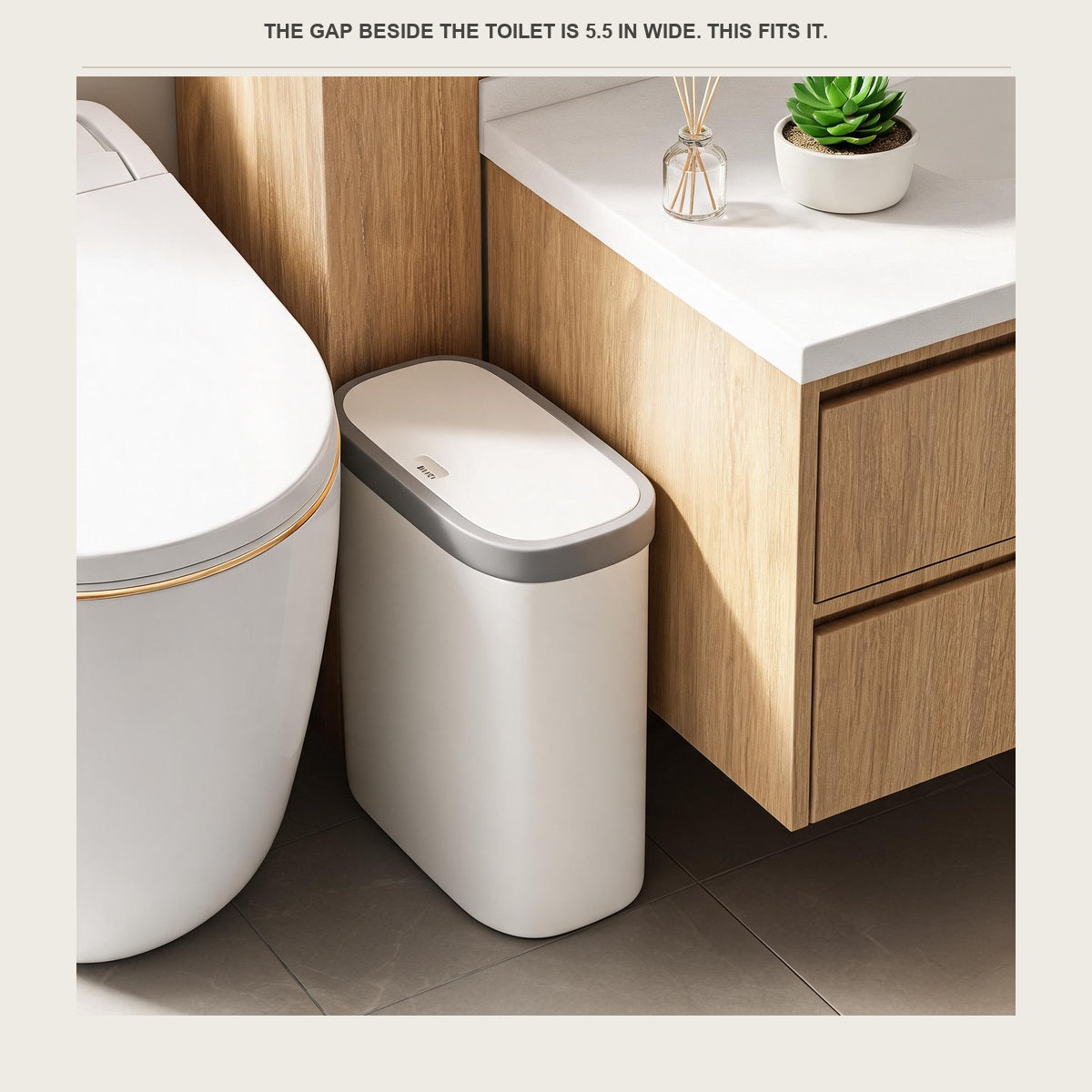 Slim white trash can with a grey rim standing in the narrow gap between a toilet and a wood vanity