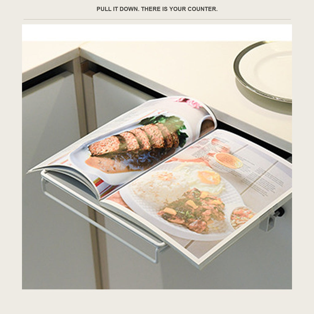 A cookbook open on the fold-down shelf clipped to a kitchen cabinet door