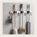Stainless steel wall mounted mop and broom holder with three cleaning tools on a tiled utility wall